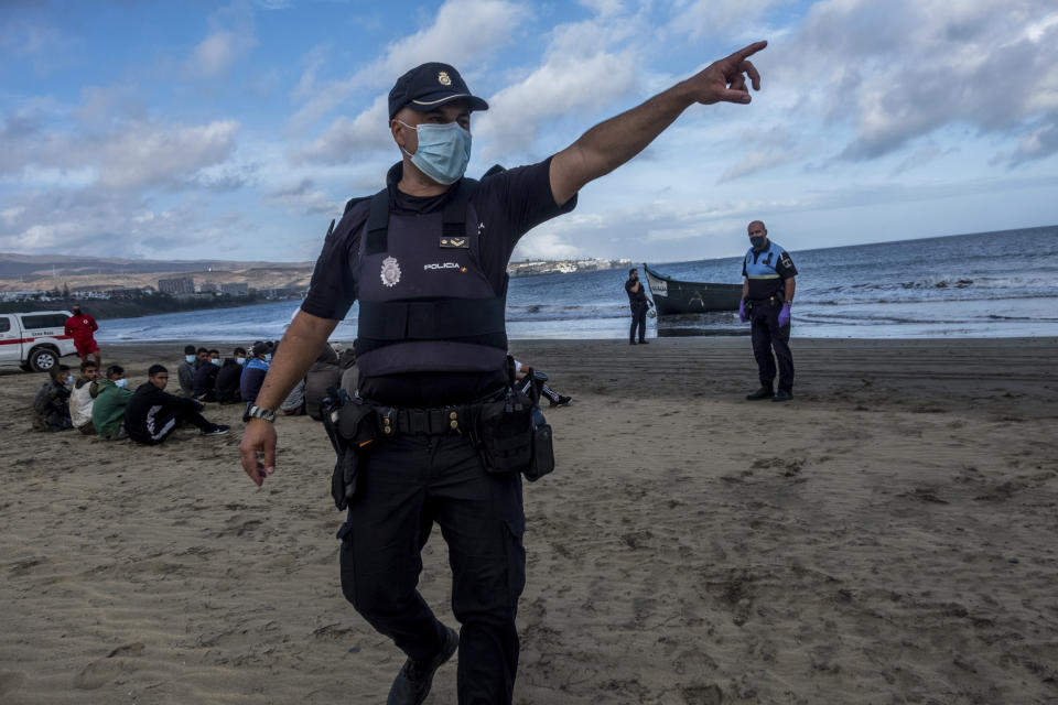 A Police officer react to the media as migrants from Morocco sit on a beach after arriving at the coast of the Canary Island, crossing the Atlantic Ocean sailing on a wooden boat on Tuesday, Oct.20, 2020. Some 1,000 migrants have spent the night again sleeping in emergency tents in a dock while authorities in the Canary Islands complain that the Spanish government keeps blocking transfers of newly arrived migrants to the mainland over coronavirus concerns. (AP Photo/Javier Bauluz)