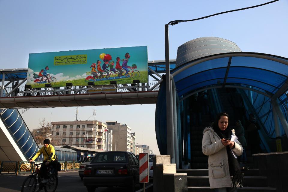 In this Tuesday, Dec. 24, 2013 photo, a woman stands while a man rides his bicycle under a banner encouraging Iranians to have more children, in a street in Tehran, Iran. Having curbed birth rates for two decades, Iran is once again promoting a baby boom to help make up for its graying population. But experts say it is difficult to encourage Iranians to have more children under a sanctions-hit economy and 36 percent inflation. (AP Photo/Ebrahim Noroozi)