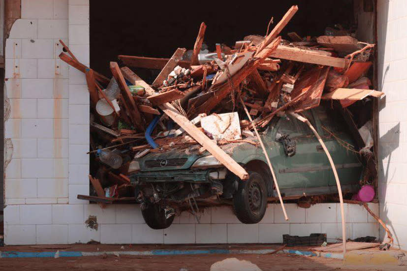 A car covered with splintered wood and debris is seen coming out of an opening in a brick wall.