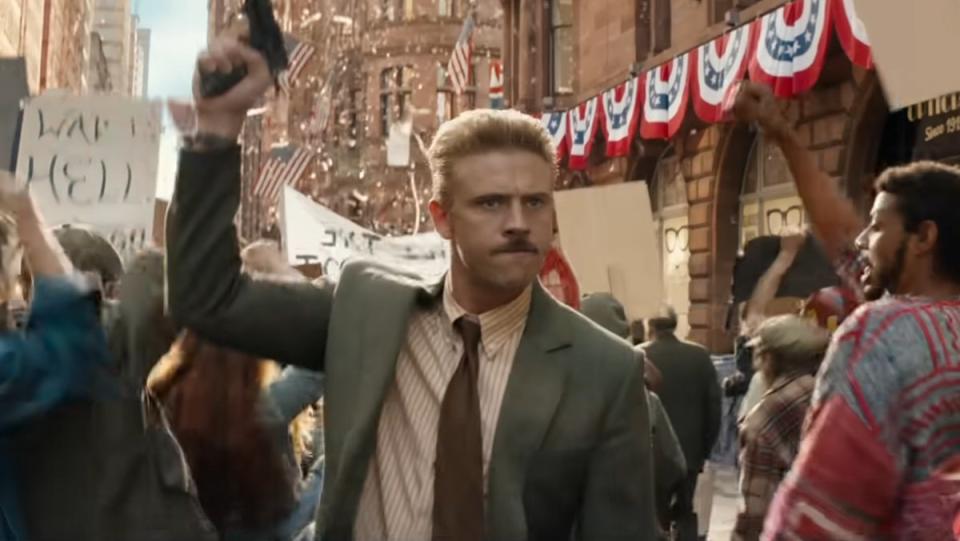 A mustachioed man with a blond crew cut in a jacket and tie holds up a gun in a parade  in Indiana Jones and the Dial of Destiny