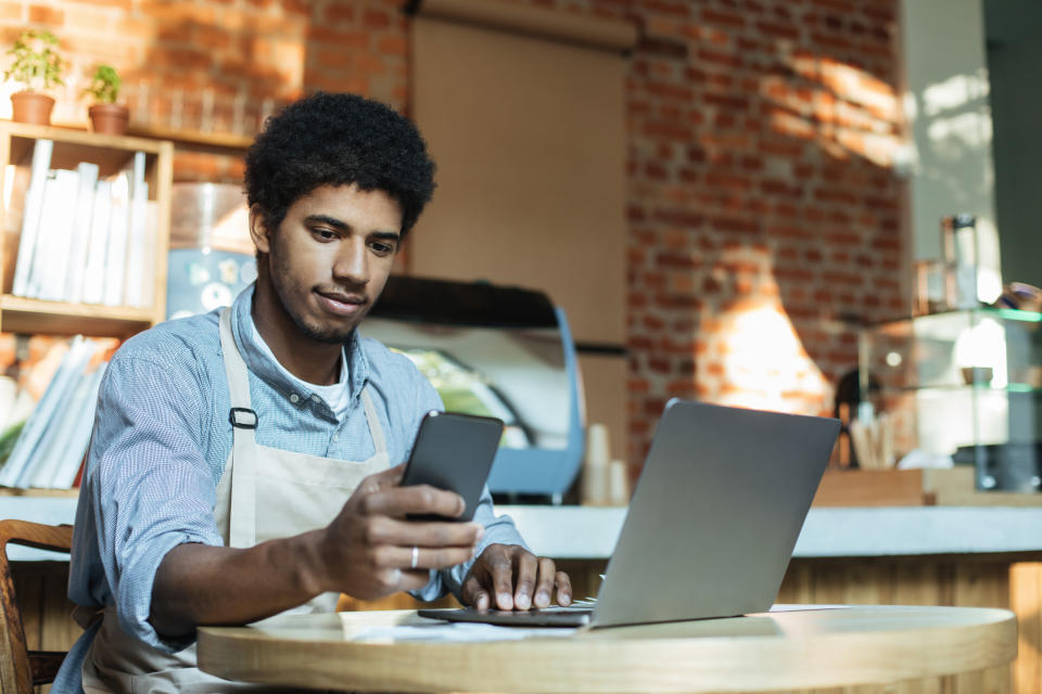 Social networking, devices and small business during covid-19 pandemic. Busy african american man in apron sits at table and works in smartphone and laptop in interior of loft coffee shop, copy space