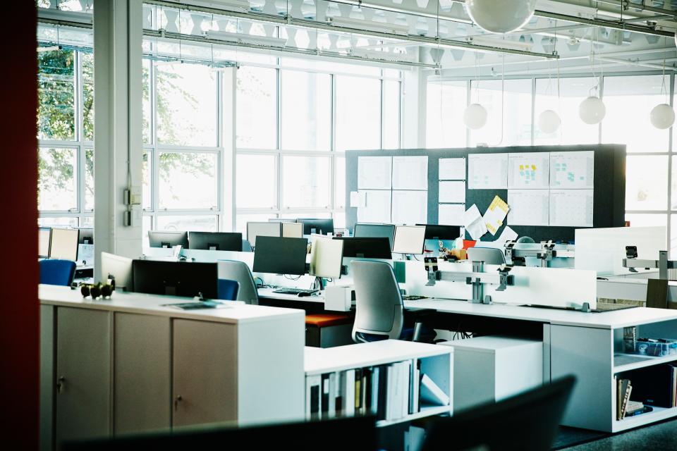 An empty office space, with computers on desks, a pinboard, and large windows.