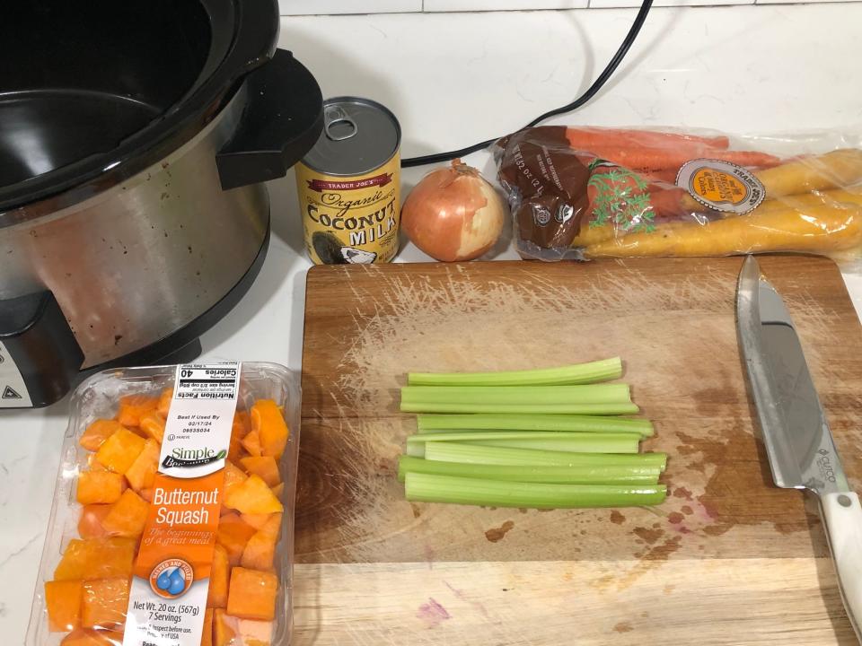 A Crock-Pot, a container of butternut squash, a can of coconut milk, a yellow onion, a bag of carrots, and a cutting board on a white counter. On the cutting board is a large knife and celery.