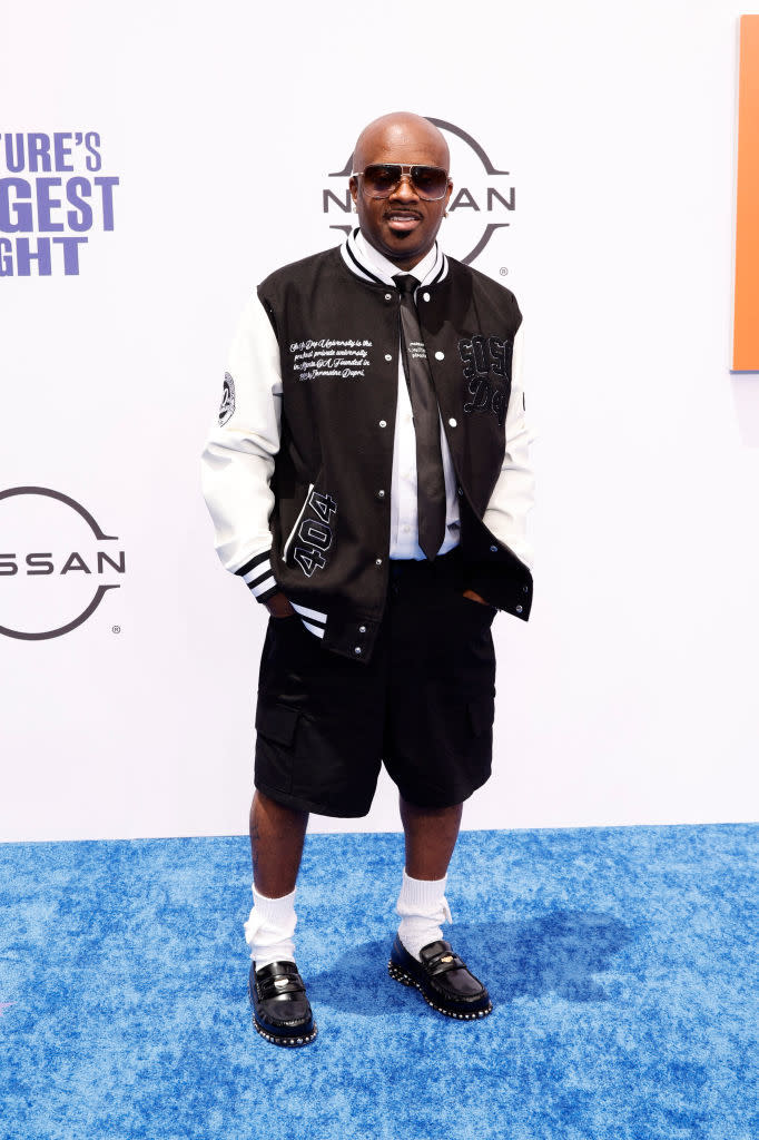 Jermaine Dupri on the blue carpet, wearing a black and white varsity jacket, black shorts, white socks, and black studded shoes