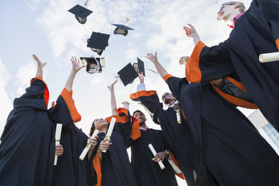 A group of seven multi-ethnic high school or university graduates wearing graduation gowns, holding diplomas. Throwing their caps in the air.