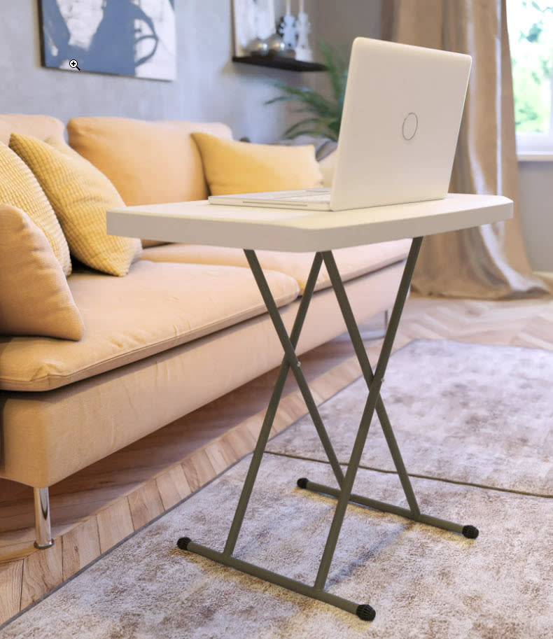 A laptop on a small, standing desk in a cozy room with a couch and cushions