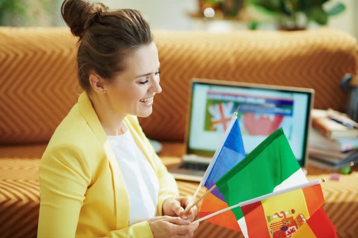 A person smiling while holding flags from three different countries, with an open laptop computer in the background.