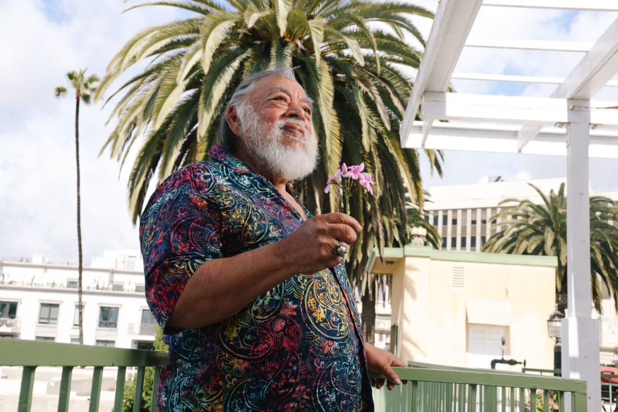 Sergio Calderon standing against a green fence wearing a patterned shirt and holding a pink flower