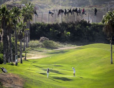 A golfer hits a tee shot as African migrants sit atop a border fence during an attempt to cross into Spanish territories between Morocco and Spain's north African enclave of Melilla in this October 22, 2014 file photo. REUTERS/Jose Palazon/Files