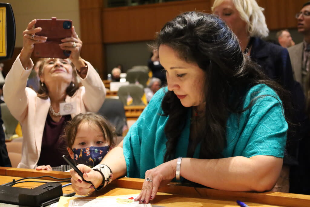 Rep. Linda Serrato in a turquoise shirt leans on a table while holding a pen to sign an official roster of state representatives in Santa Fe, New Mexico. Her young daughter peeks ahead, she is wearing a mask. Behind her is Serrato's mother in a white blazer holding a cellphone above her head to take a picture.