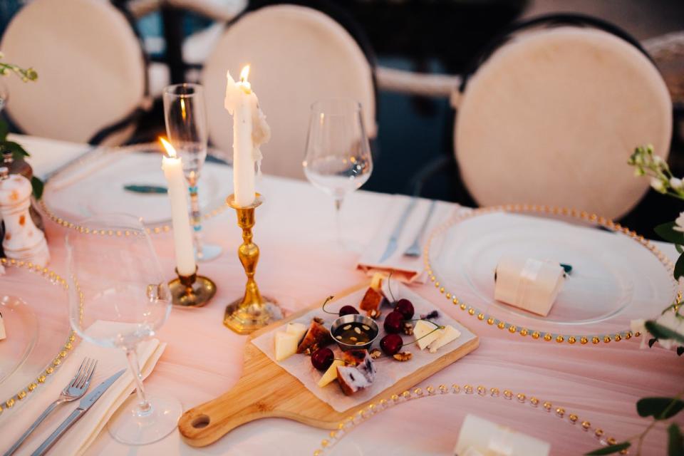 Table decorated with plates with beads and candles on a white tablecloth and cherries and cheese on a cutting board. 