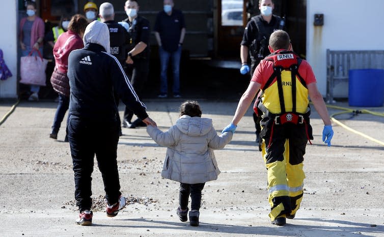 View from behind of a young child walking between an adult man and an RNLI rescue crew member, each holding one of the girl's hands