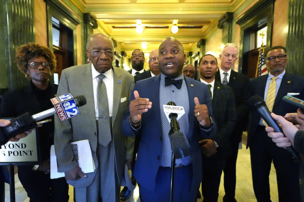 A man in a blue suit, blue shirt and bow tie surrounded by legislators and speaking to a stand of microphones