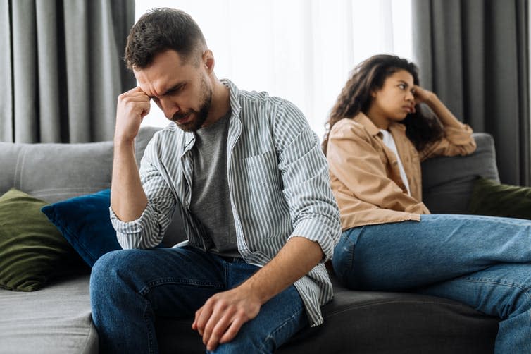 A young couple sit on a couch facing opposite directions, both looking frustrated.