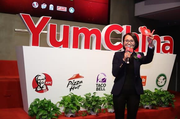 Yum China CEO Joey Wat speaking in front of a company sign with the KFC, Pizza Hut, and Taco Bell logos displayed.