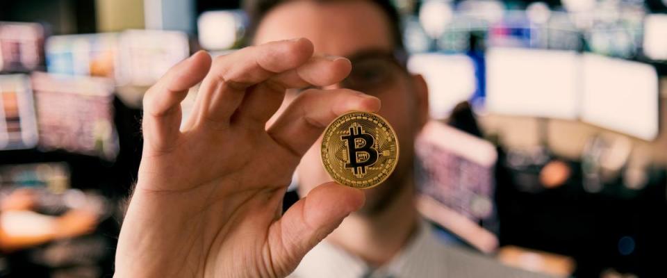 Man stands in office holding up bitcoin between thumb and forefinger to the camera.