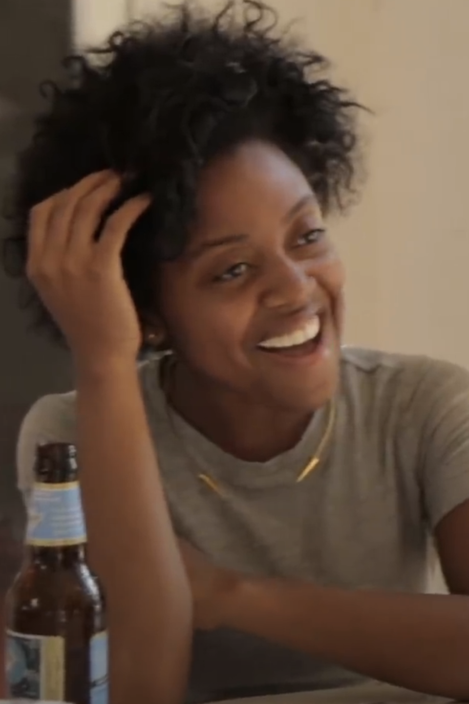 A person with curly hair, wearing a grey t-shirt, smiling and sitting at a table with a bottle of beer in front of them