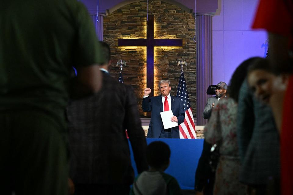 A man in a dark suit and red tie, raising his fist in a church against the background of a large cross and two US flags.