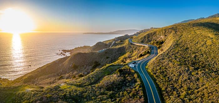 Aerial view of a curvy coastal road with a car, sea on the right and hills on the left at sunset