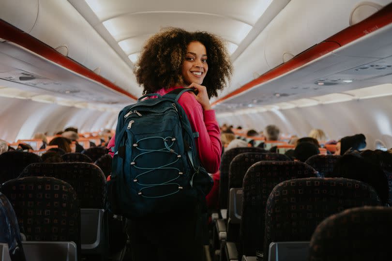 A young woman boards a small commercial airplane, carrying a backpack. She walks down the middle aisle, glances over her shoulder, and smiles at the camera.