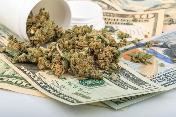 A tipped-over cannabis bottle with dried buds atop a messy pile of dollar bills.