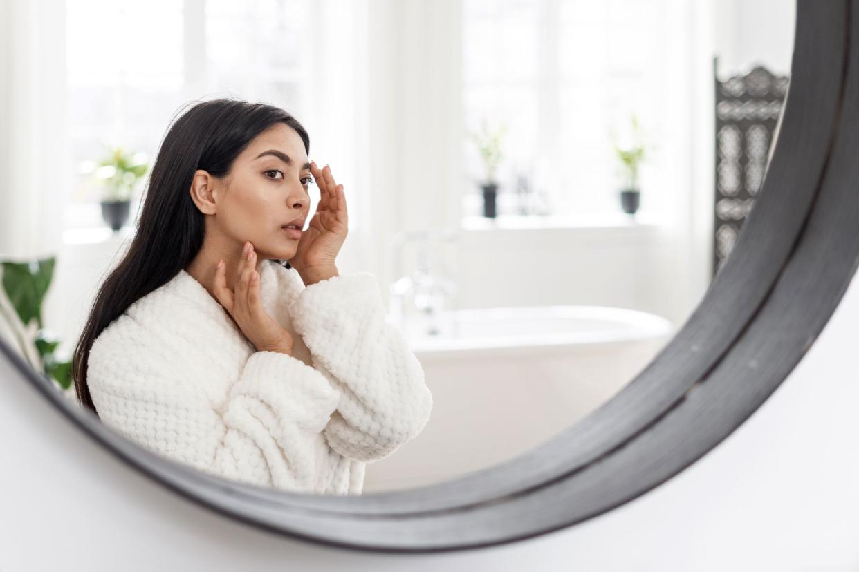 Round mirror reflection of young woman wearing a white robe, inspecting her face with bath tub, windows of bathroom in the background, mirror is rimmed with black, on a white wall