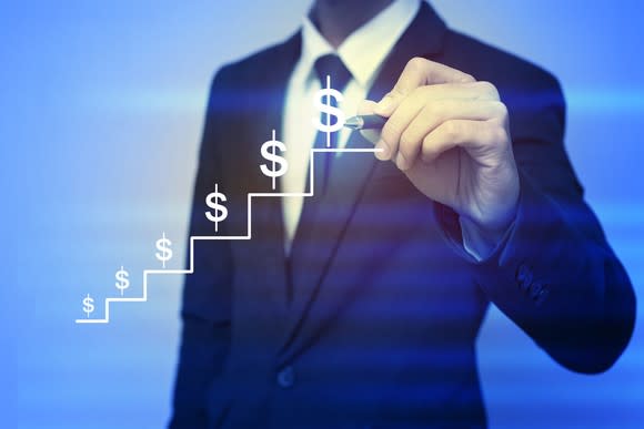 A man in a suit holds a pen to a transparent drawing of ascending steps with dollar symbols on them.