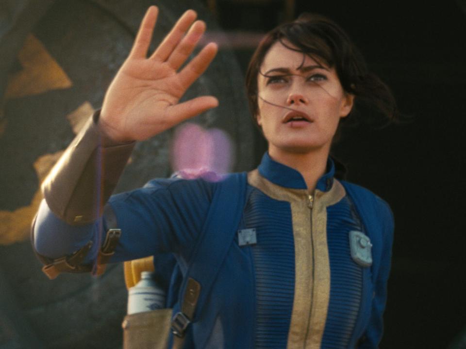 ella purnell as lucy in fallout, pushing aside a panel and wearing a blue and yellow dirty jumpsuit