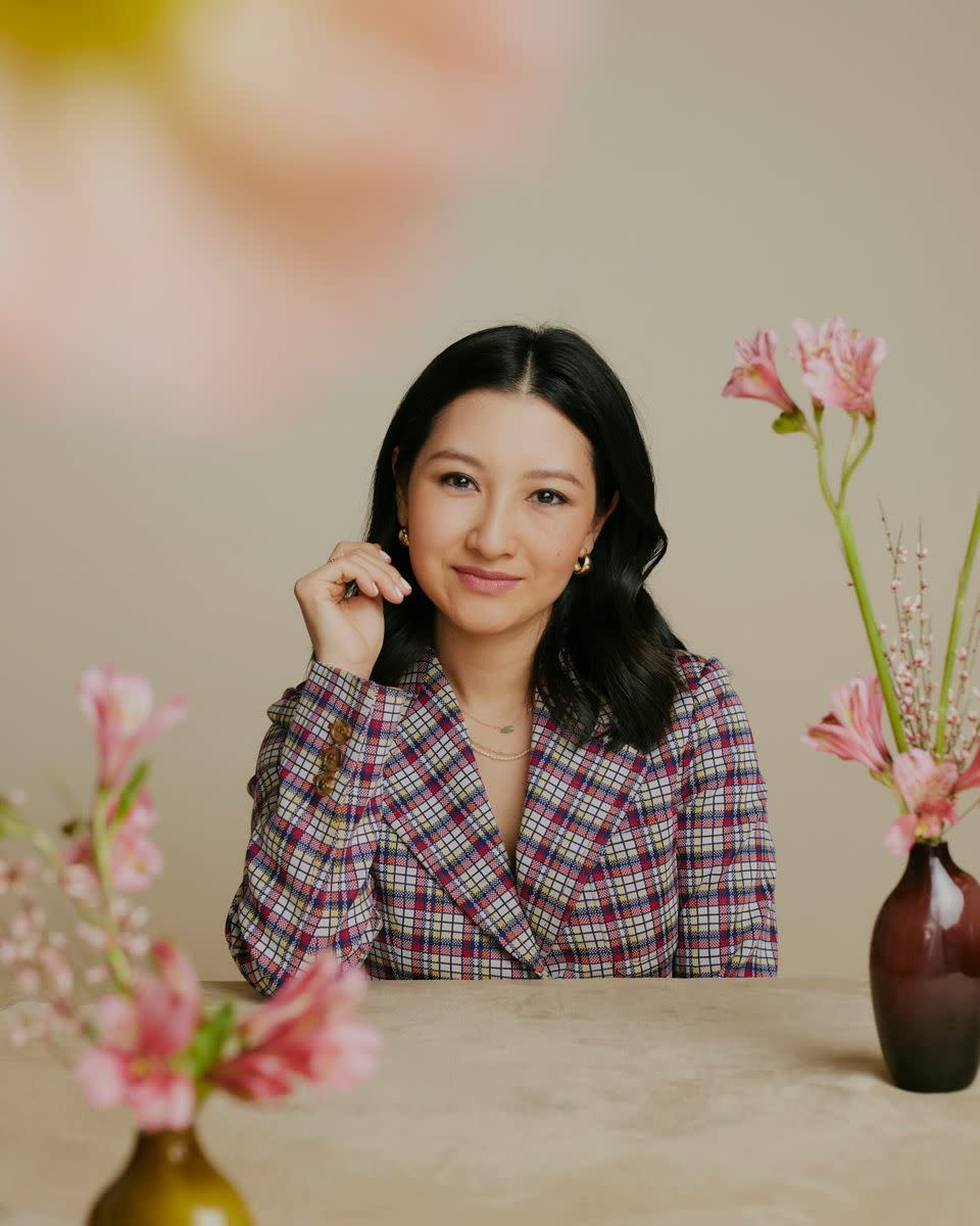 designer crystal ung of ren jewelry wears a red, yellow, blue and white plaid blazer and sits at a table with several vases of pink flowers