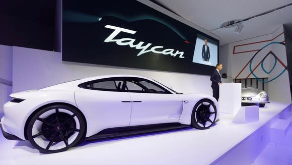 Blume is standing on an auto-show stage with the Taycan, a sleek white sports sedan, and a vintage Porsche sports car.