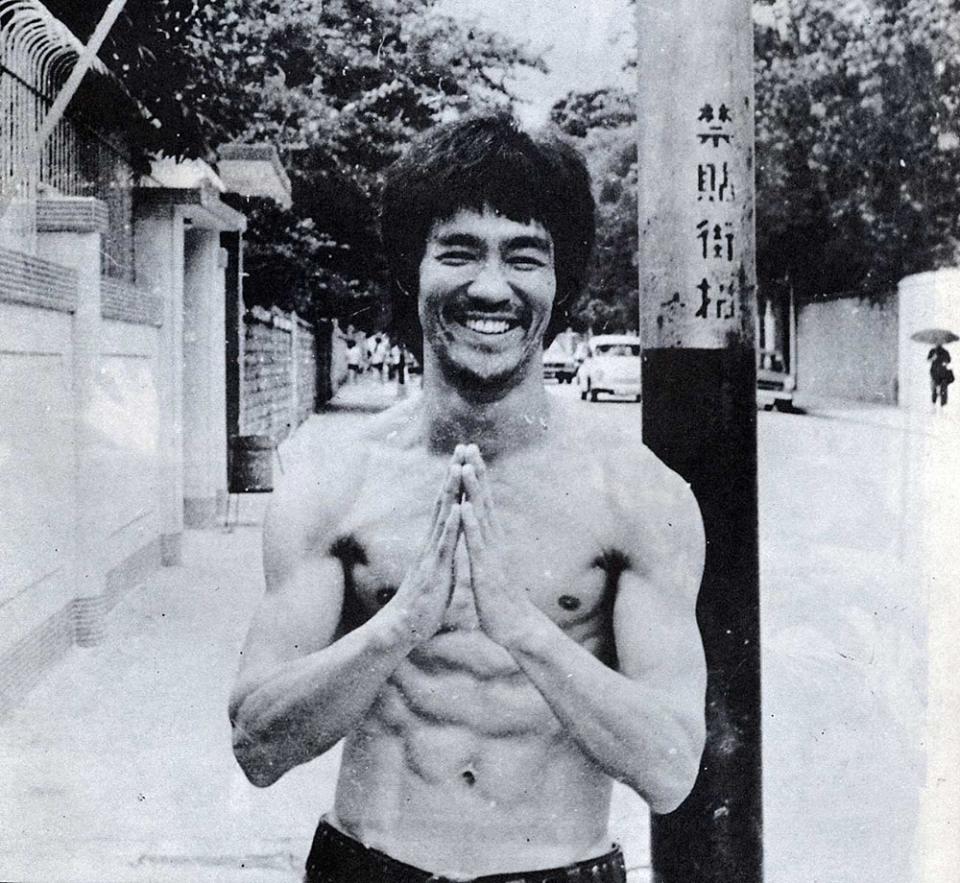 Bruce shirtless, smiling, hands together, pray motion, beard, standing in the street.