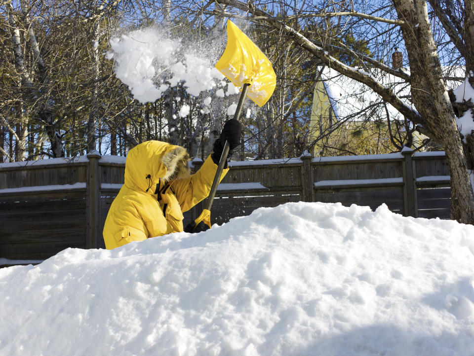 A man in a yellow parka shovels snow from a walkway after a snow storm.   Snow is chest deep and the shovel has snow flying in the air.  Country rural scene with copy space in the snow image bottom.