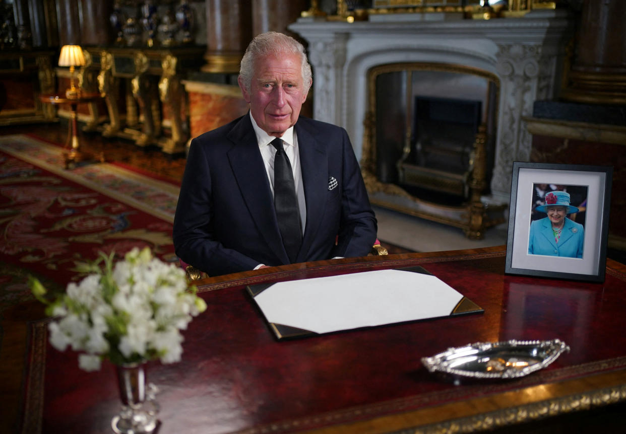 King Charles III sits at a desk on which rest flowers and a picture of Queen Elizabeth.