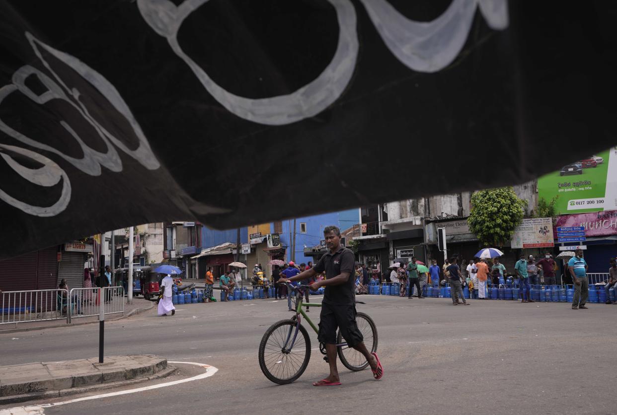 Sri Lankan man pushes his bicycle as people demanding for cooking gas sit with their empty gas cylinders blocking a busy intersection for the second consecutive day in Colombo, Sri Lanka, Sunday, May 8, 2022. Diplomats and rights groups expressed concern Saturday after Sri Lankan President Gotabaya Rajapaksa declared a state of emergency and police used force against peaceful protesters amid the country's worst economic crisis in recent memory. (AP Photo/Eranga Jayawardena)