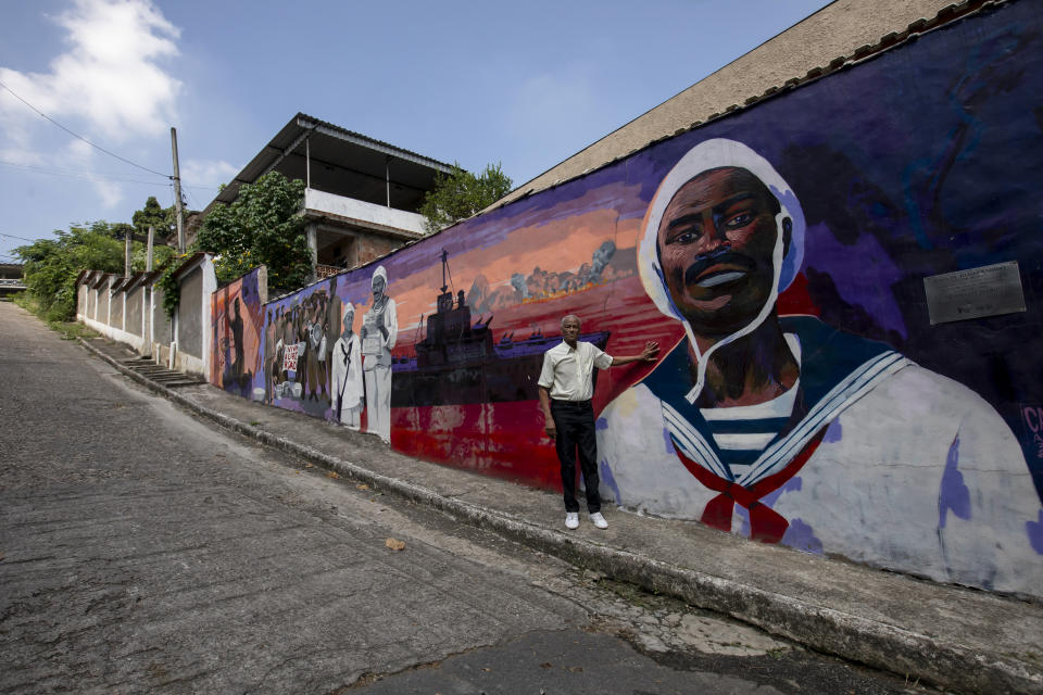 Adalberto Cândido poses for a photo in front of a mural that depicts the story of his father João Cândido, a black sailor who led a revolt against the Brazilian Navy, in Sao Joao de Meriti, Rio de Janeiro state, Brazil, Thursday, Dec. 21, 2023. After witnessing a sailor's flogging, Cândido led a revolt against regular whipping by officers in 1910. He and fellow mutineers were tortured, and only two survived — including Cândido. (AP Photo/Bruna Prado)