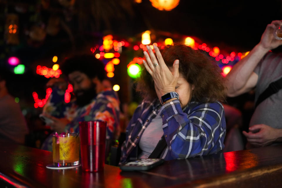 A woman in a plaid shirt sits at a bar with a hand over her face, while a man in a colorful shirt stands in the background
