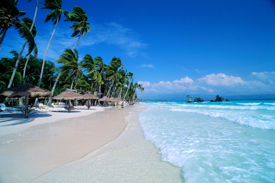  Boracay Island in the Philippines, photo shows stretch of white sand beach, gentle waves, palm trees and thatched huts