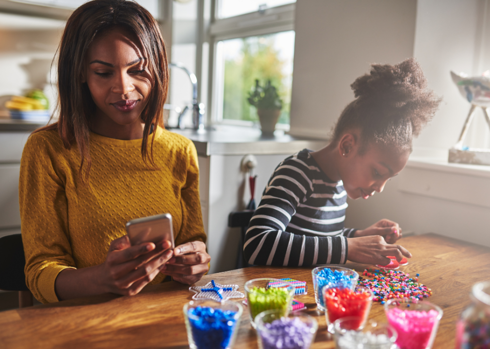 A woman checking her phone while a little girl does crafts next to her.