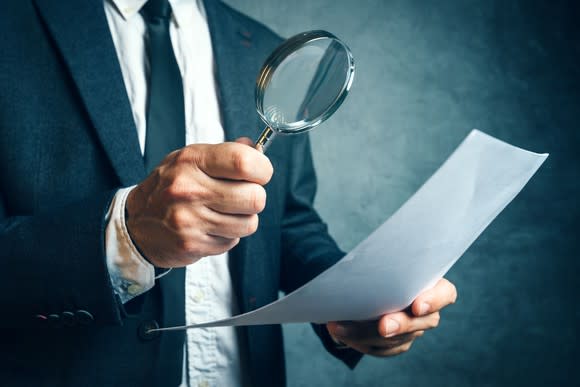 man in a suit looks at a piece of paper through a magnifying glass.