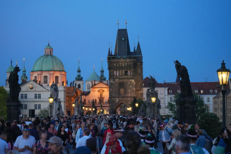 A bustling crowd on the Charles Bridge in Prague, with statues, historic towers and church domes in the background at dusk