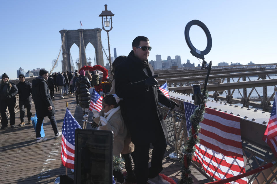 Tourists take a video with the help of a vendor working on the Brooklyn Bridge in New York, Tuesday, Jan. 2, 2024. New York City will ban vendors from the Brooklyn Bridge starting Wednesday, Jan. 3, 2024. The move is intended to ease overcrowding on the famed East River crossing, where dozens of souvenir sellers currently compete for space with tourists and city commuters. (AP Photo/Seth Wenig)