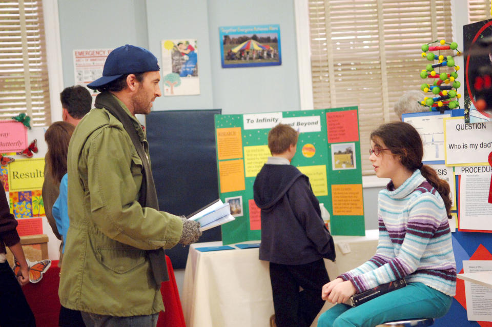 Scott Patterson, wearing a jacket and hat, talks to Alexis Bledel, dressed in a sweater, at a school science fair featuring various project displays in the background