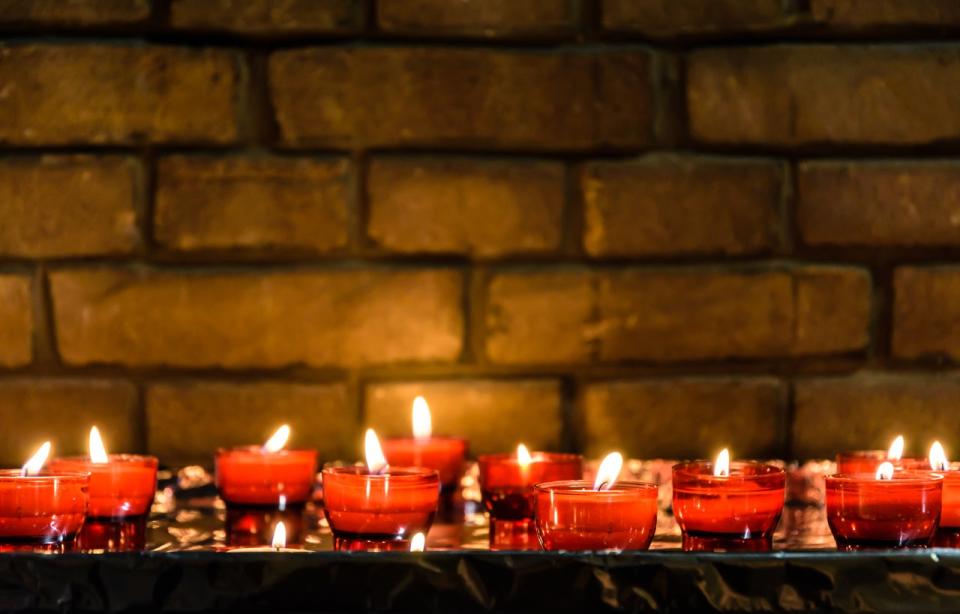 Small, lit white votive candles in red glass holders on a table covered with foil in front of a brick wall.