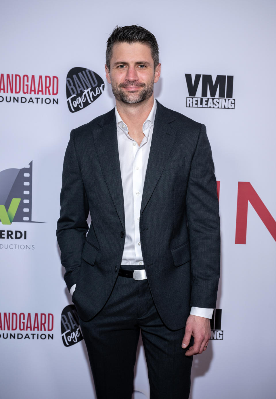 A man in a tailored suit poses on a red carpet in front of a backdrop with logos