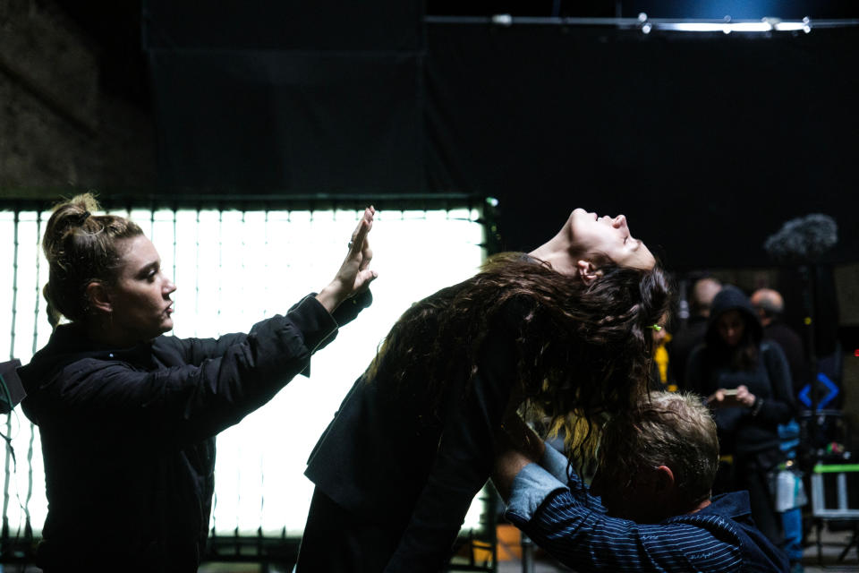 Two people style a woman's hair on a film set, with lighting equipment in the background