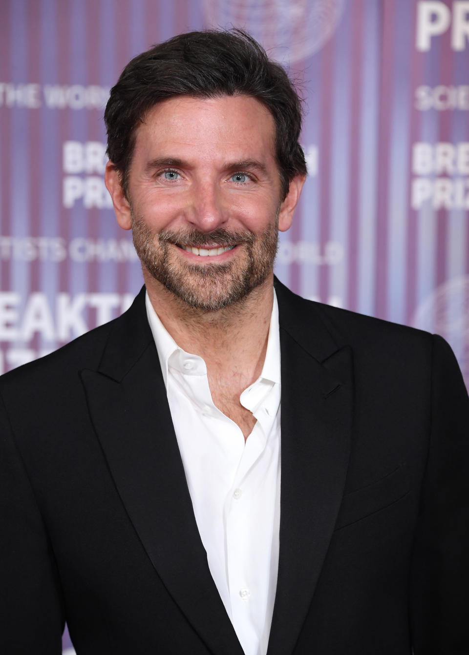 Man in a black suit and white shirt smiling at a camera on a purple backdrop