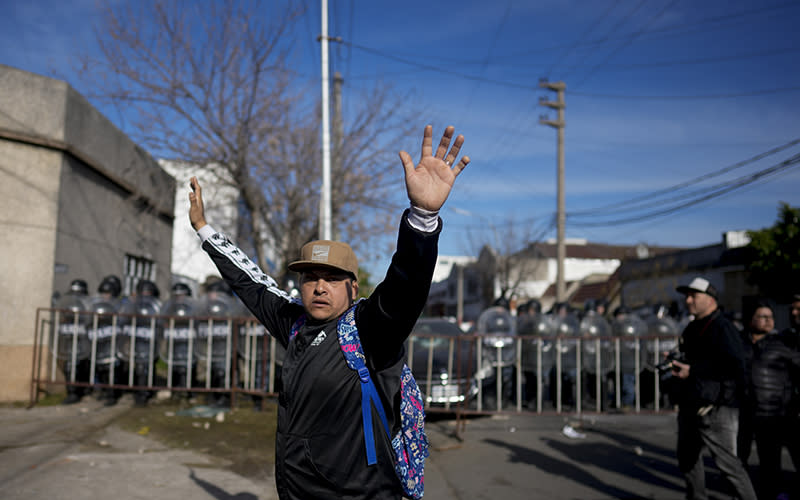 A man raises his arms during a protest. Behind him is a line of police wearing riot gear and carrying shields