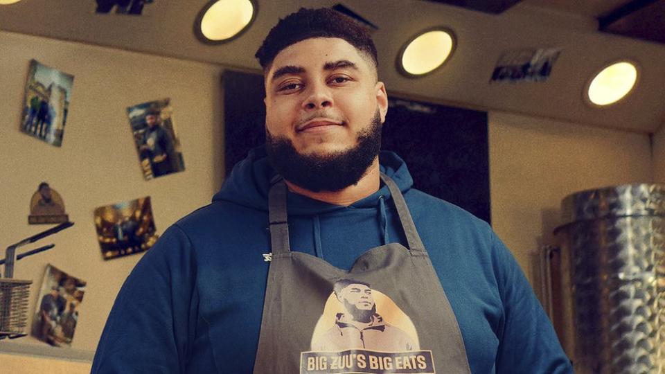 Big Zuu from his BBC series Big Zuu's Big Eats. Big Zuu is a 28-year-old black man with short afro hair, brown eyes and a dark beard. He wears a grey apron with 'Big Zuu's Big Eats' printed on it over a blue hoodie. He's pictured inside a kitchen which has photos printed on the walls.