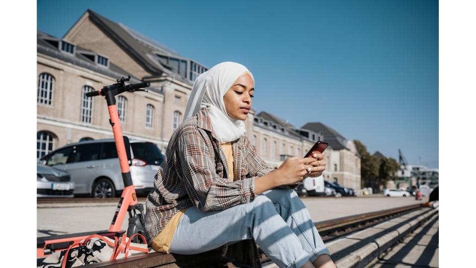 young muslim woman with hijab sitting at riverside in berlin and tapping at  mobile