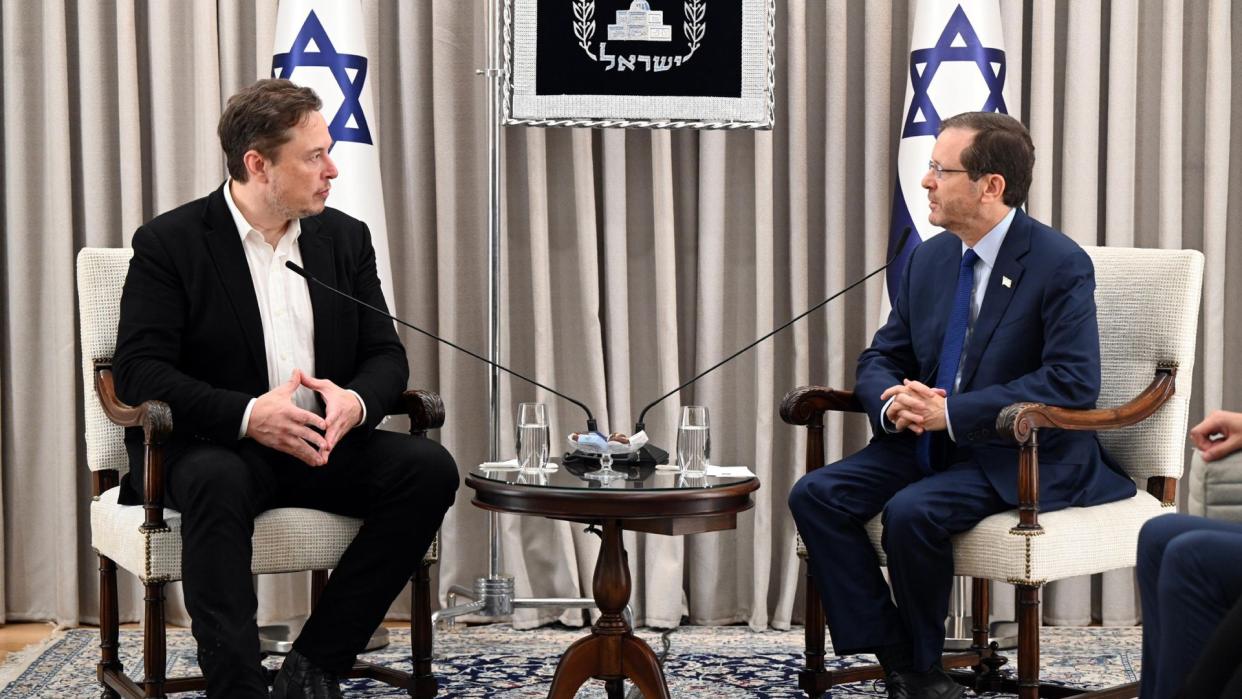  Two men in suits speak to one another in front of microphones and Israeli flags. 